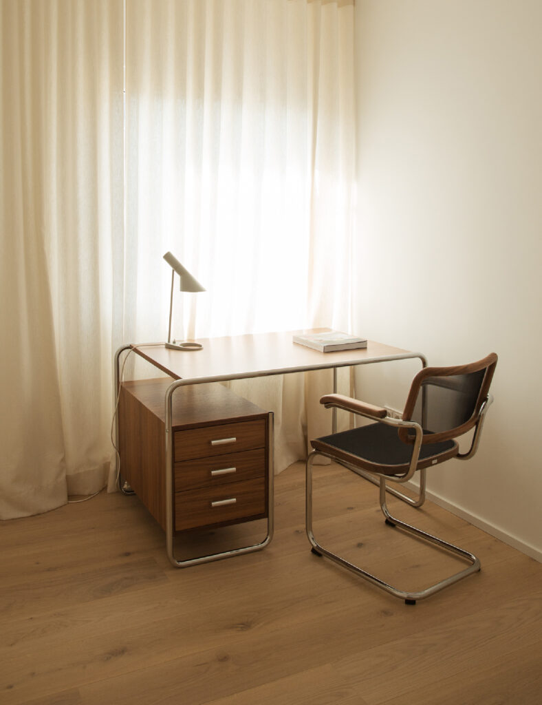 Ein Mietstudio mit einem Schreibtisch und einem Stuhl vor einem Fenster, das Tageslicht hereinlässt.